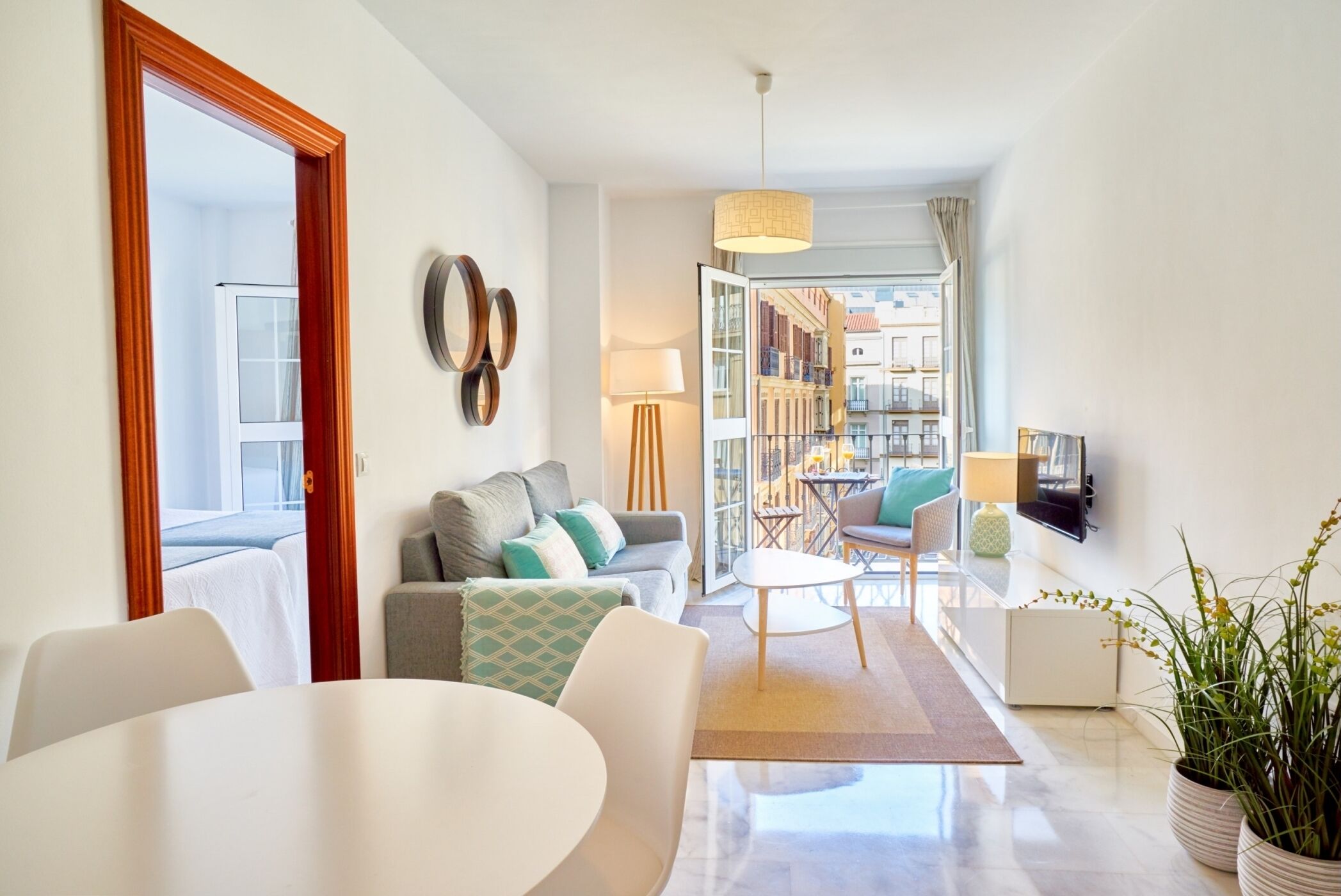 A modern living room with a sofa, coffee table, and chairs. There is a TV mounted on the wall and a window providing a view of the city.