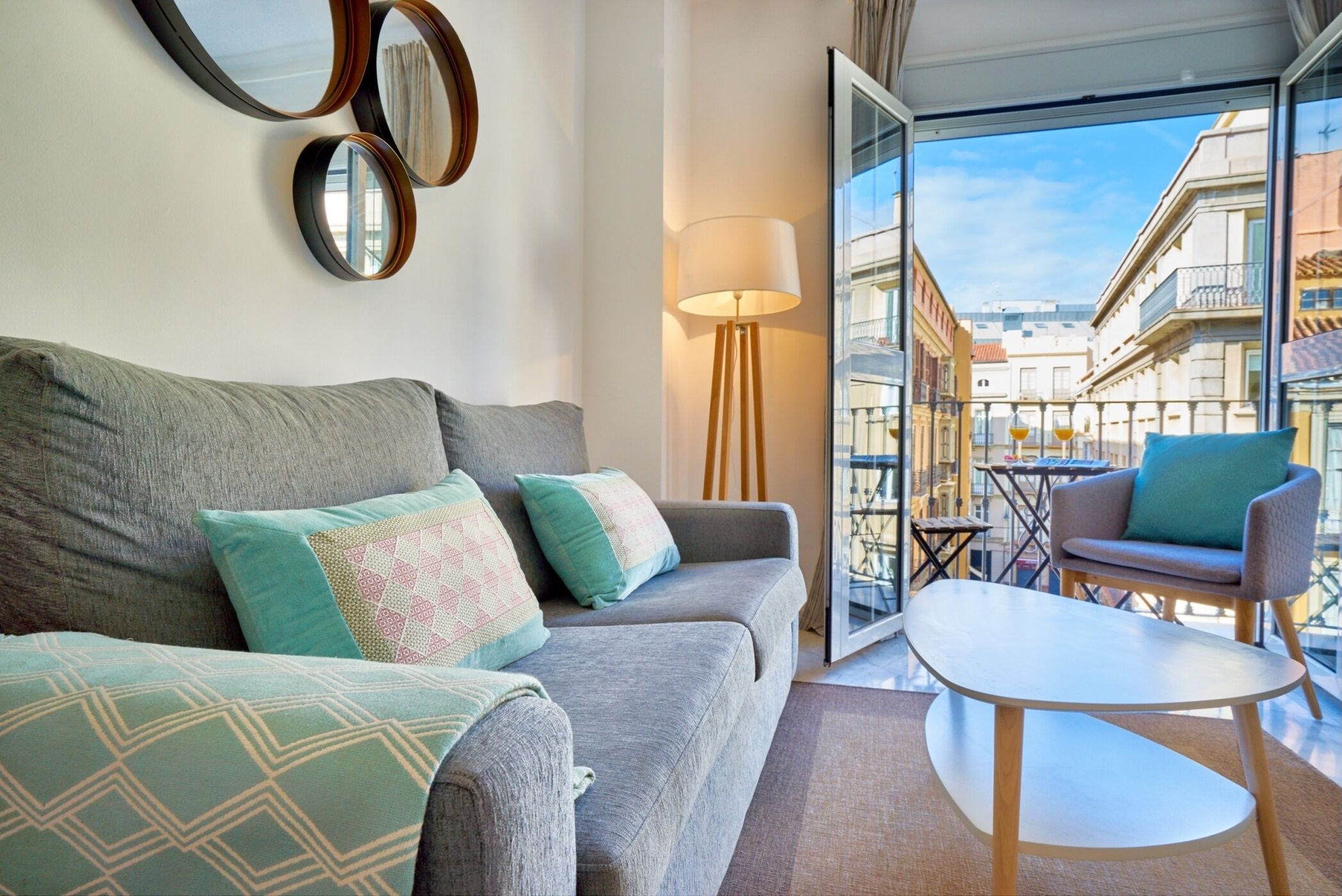 A modern living room with a grey sofa, patterned cushions, a round table, and a floor lamp. A large window offers a view of a cityscape.