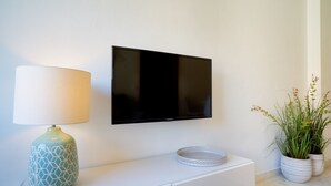 A flat-screen TV mounted on a white wall, a decorative lamp with a geometric pattern, and two potted plants on a white cabinet. - iloftmalaga Puerta del Mar II (Málaga)