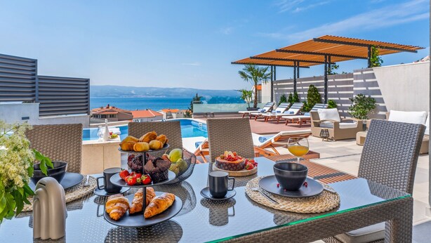 A dining table set with food and drinks, overlooking a pool and a beach.