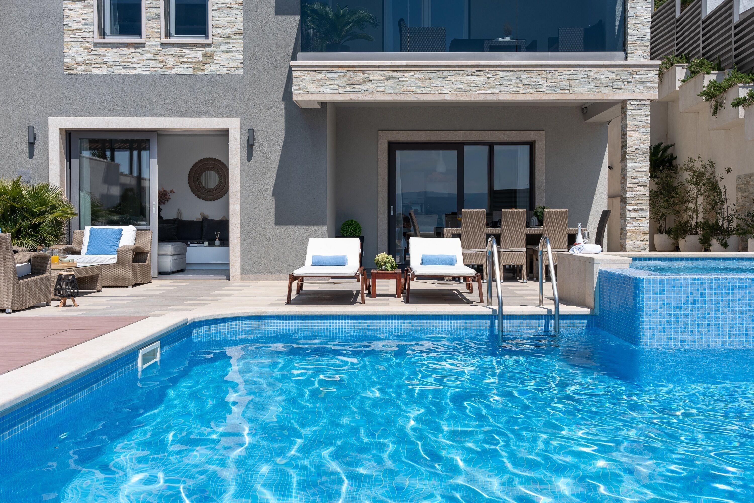 A modern outdoor swimming pool with two lounge chairs, a small table, and a glass door entrance.