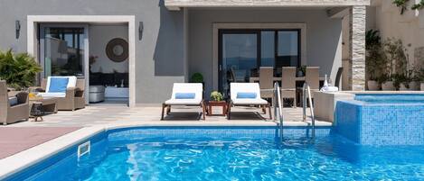 A modern outdoor swimming pool with two lounge chairs, a small table, and a glass door entrance.