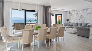 A modern living room with a dining area, large windows with a view of the sea, and a grey sofa.