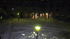 A snow-covered courtyard with a lit lamp post and buildings in the background.