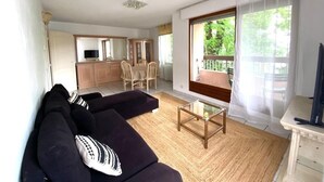 A living room with a sofa, a coffee table, and a TV. There is a kitchen area with cabinets and a dining table.