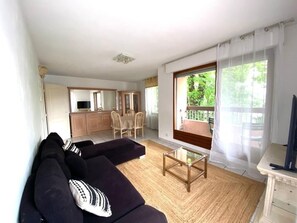 Ein Wohnzimmer mit einer Couch, einem Couchtisch und einem Fernseher. Es gibt eine Küchenzeile mit Schränken und einem Esstisch.