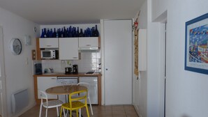 Cucina compatta con mobili bianchi, un forno a microonde e una macchina da caffè. C'è un tavolo da pranzo con due sedie, una gialla e una bianca. Un quadro incorniciato è appeso al muro, e si vede una porta sullo sfondo.