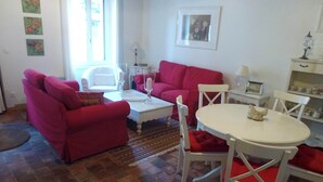 Ein Wohnzimmer mit einem roten Sofa, einem weißen Couchtisch und einem Esstisch mit Stühlen. Ein Fenster bietet einen Ausblick nach draußen.