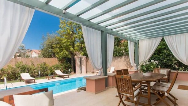 A covered outdoor patio with a pool, wooden furniture, and white curtains. - Villa Il Rifugio, villa with private pool (Pietrasanta)