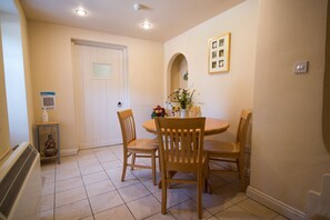 A dining area with a round table, four wooden chairs, a vase with flowers, a framed picture on the wall, and a door with a frosted glass window. - Beautifully appointed cottage in extensive grounds (Penrith)