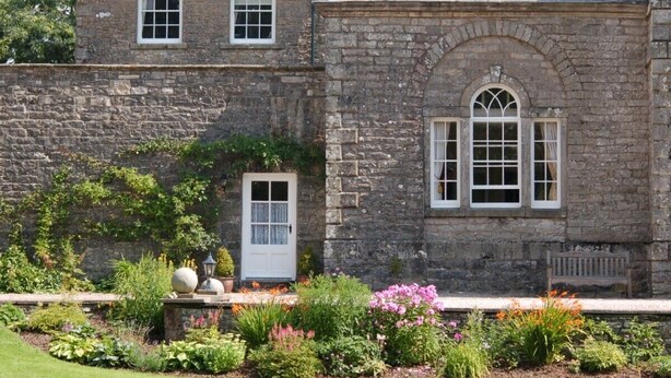 A stone building with a white door and windows, surrounded by a well-maintained garden with various plants and flowers.