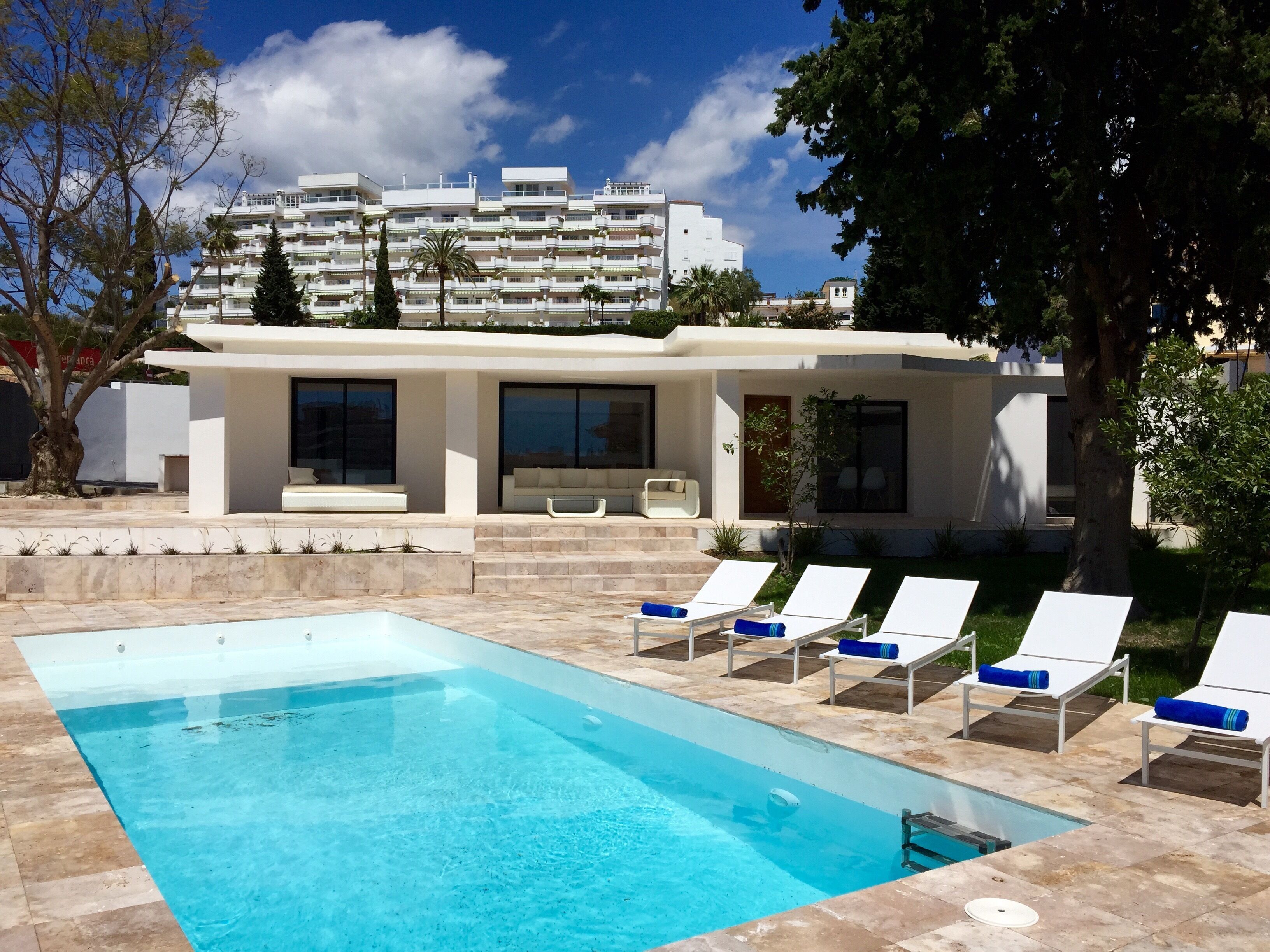 Un área de piscina moderna con una piscina cristalina, sillas de descanso blancas y un edificio con grandes ventanales al fondo.
