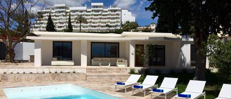 Un área de piscina moderna con una piscina cristalina, sillas de descanso blancas y un edificio con grandes ventanales al fondo.