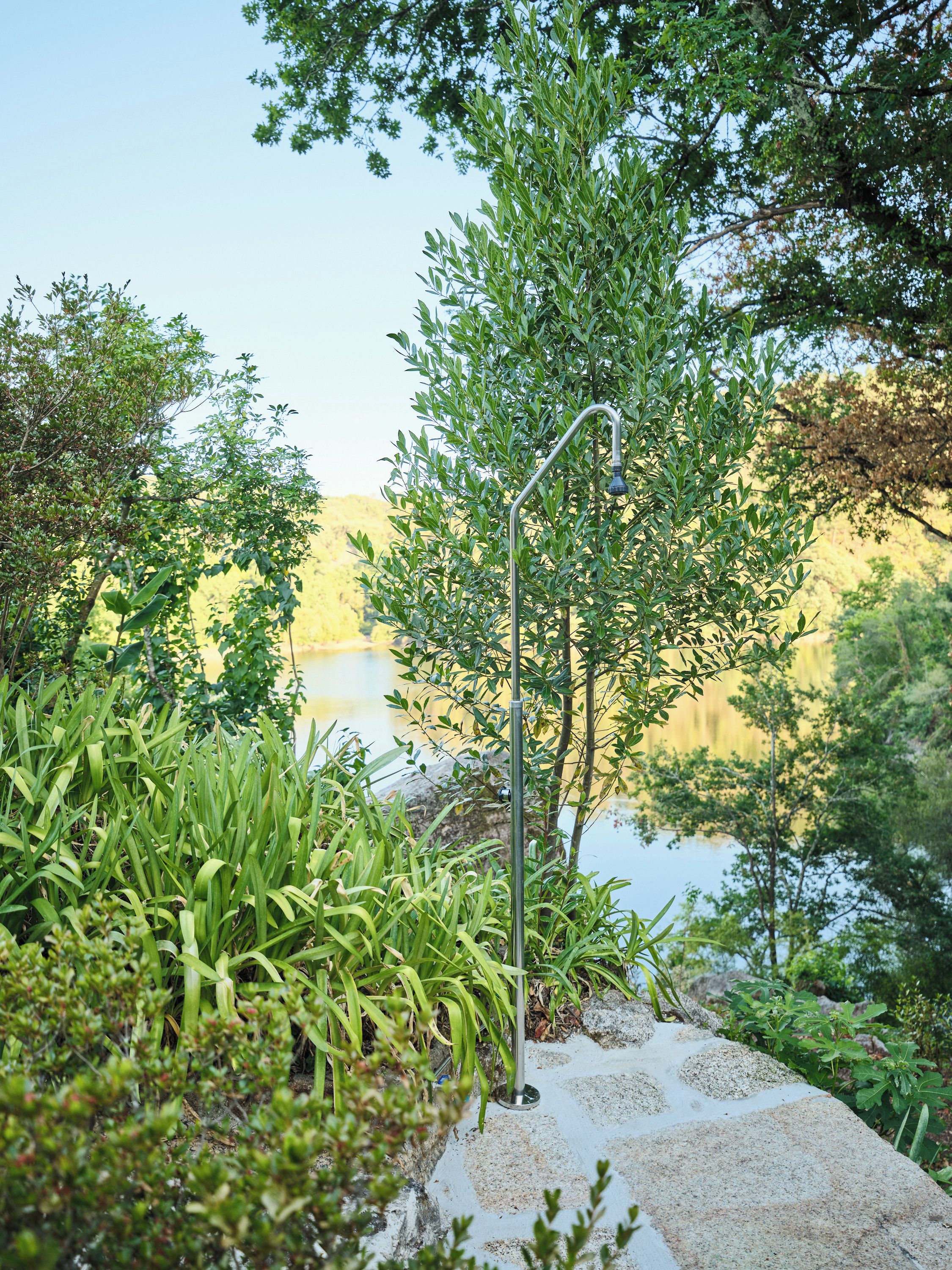 Ein Garten mit einer Metallkonstruktion, möglicherweise einem Laternenpfahl, umgeben von viel Grün und einem Gewässer im Hintergrund.