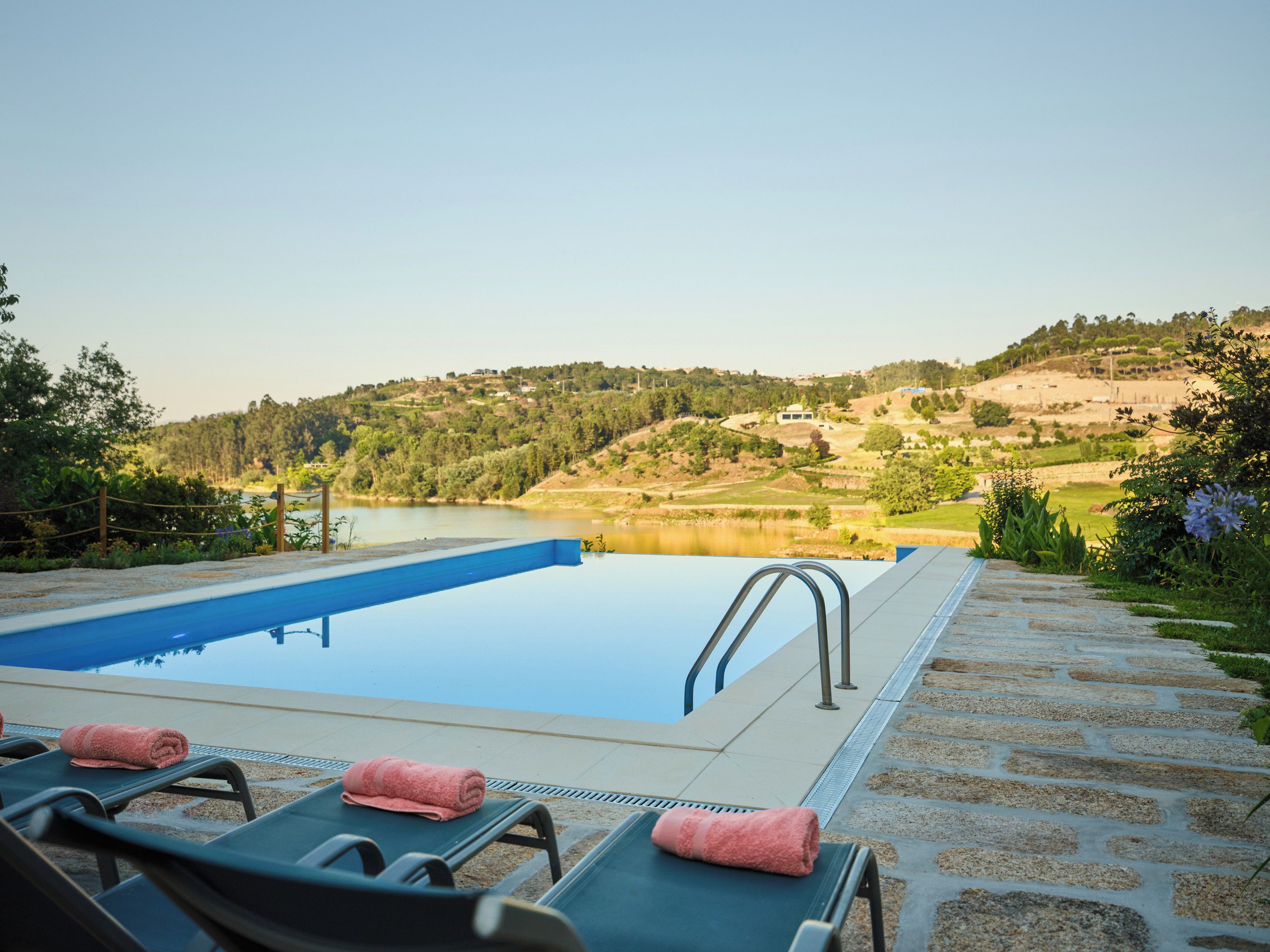 Piscina con sillones de descanso y una toalla, con vista a un lago y un paisaje montañoso.