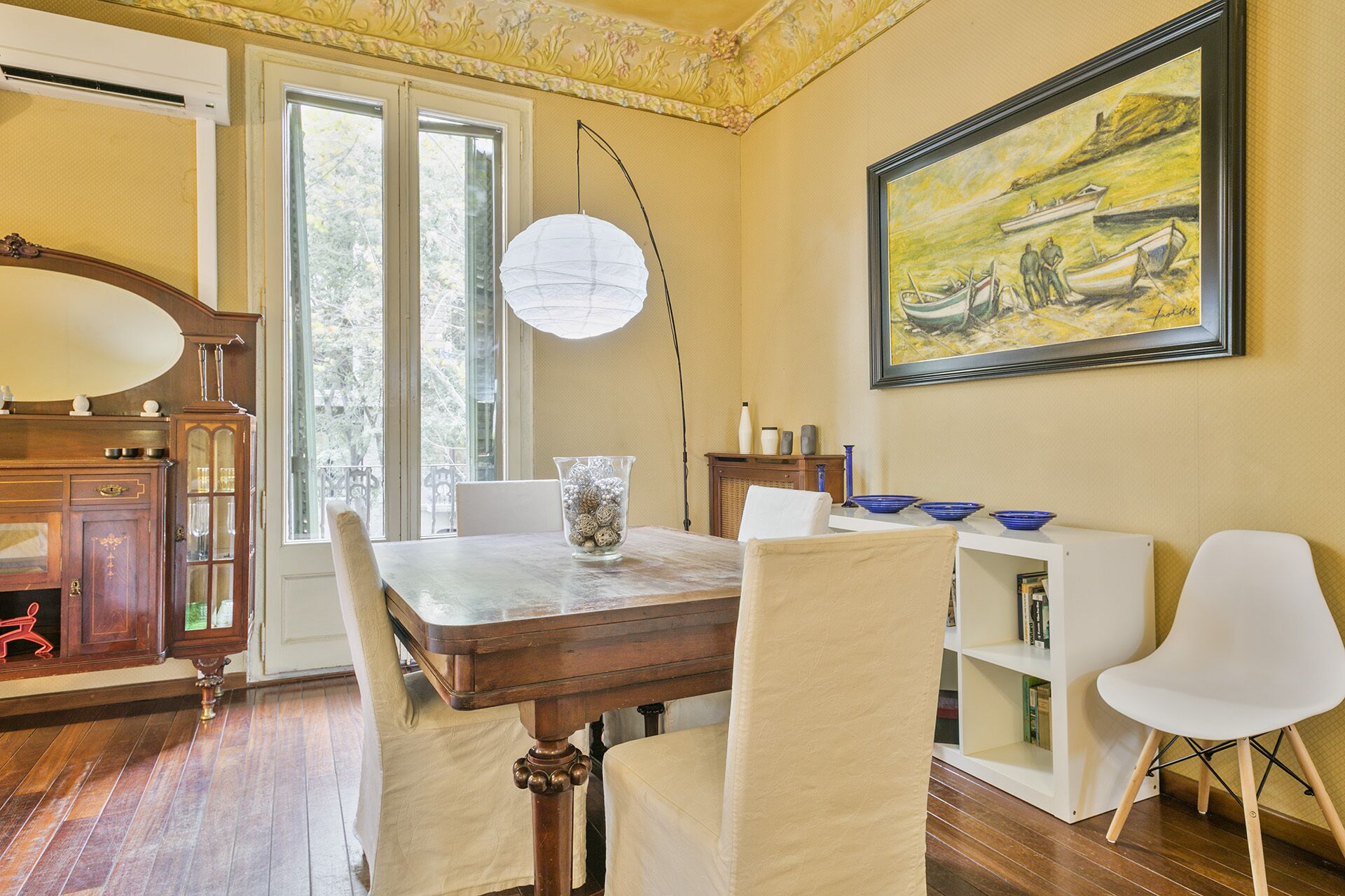 A dining area with a wooden table, white chairs, a vase, and a painting on the wall.