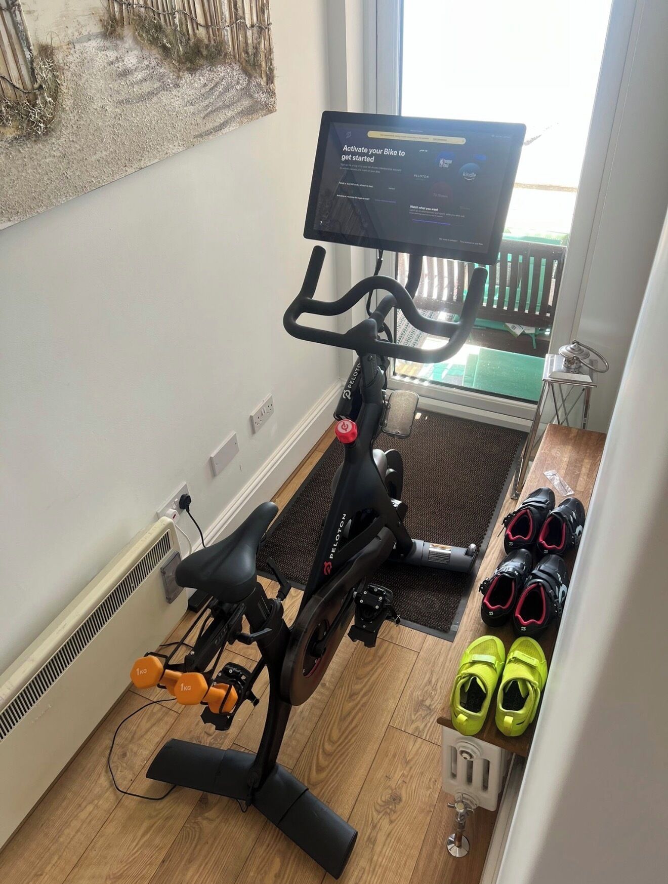 A Peloton stationary bike with a screen, a mat, and a pair of shoes on the floor.