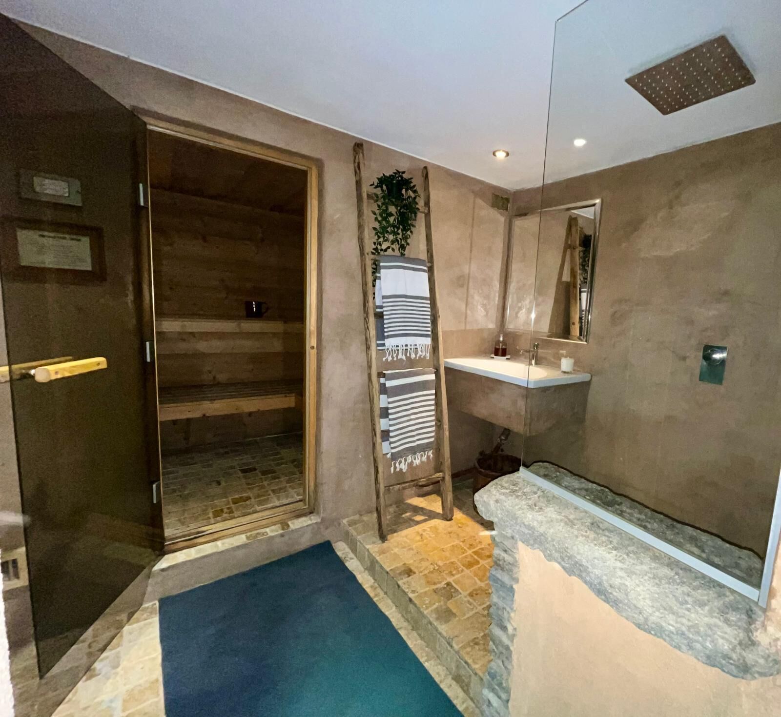 Una habitación con puerta de madera, un tapete azul, un banco de piedra, un lavabo y un espejo.