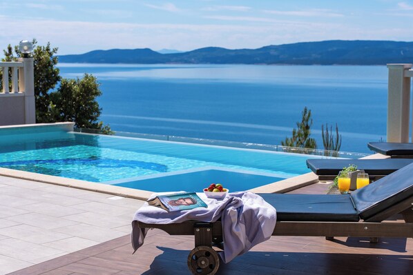 A poolside area with a view of the sea, a lounge chair, a table with a book and fruit, and a towel. - Villa MIRNA with heated pool & whirlpool, traditional wine bar, 150m from sea (Lokva Rogoznica)