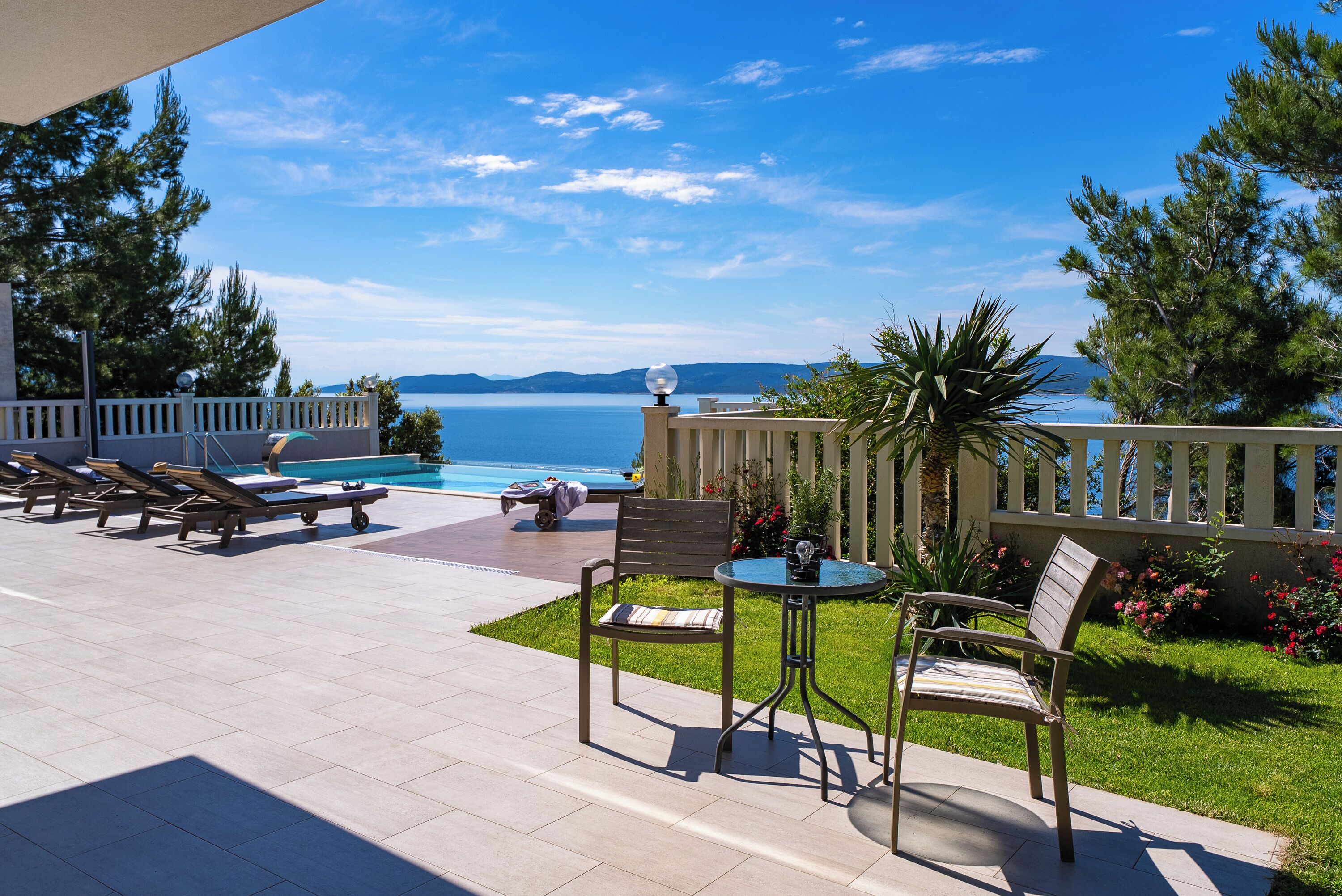 A poolside area with lounge chairs, a table, and a chair, overlooking a body of water with mountains in the distance.