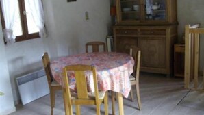Un pequeño comedor con una mesa redonda cubierta con un mantel floral, sillas de madera, un aparador con puertas de cristal y una ventana con cortinas.