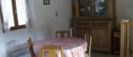 A small dining area with a round table covered by a floral tablecloth, wooden chairs, a cabinet with glass doors, and a window with curtains.