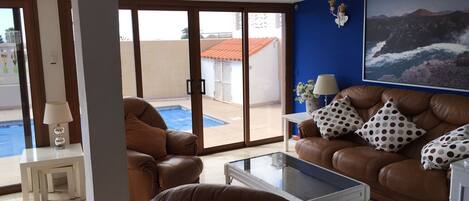 Un salón con un sofá de cuero marrón, una mesa de centro de vidrio y vistas a una piscina a través de puertas de cristal.