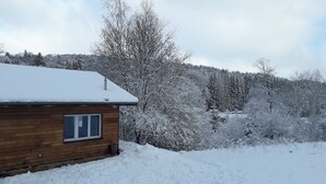 Une cabane en bois avec une fenêtre, entourée d’arbres couverts de neige et avec une colline en arrière-plan.