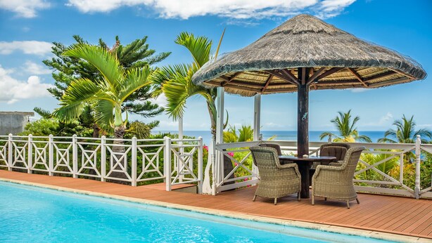 Un área junto a la piscina con un quiosco de paja, muebles de mimbre y vista al océano.