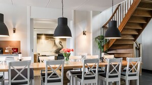 Uma sala de jantar moderna com mesa de madeira e cadeiras brancas, luminárias pendentes pretas e uma escadaria.