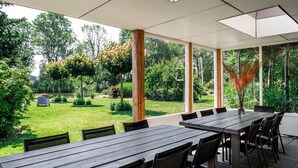 Um pátio externo com uma longa mesa de madeira e cadeiras, cercado por amplas janelas que dão para um jardim bem cuidado.