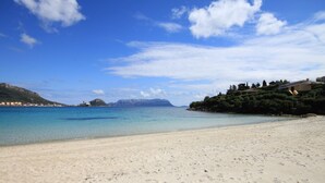 A beach with clear water, sandy shore, and buildings on the right. - Villaggio Perlacea - PERLACEA 32 (Golfo Aranci)