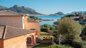 A coastal building with a tiled roof, a stone archway, and a view of the sea and mountains. - Villaggio Perlacea - PERLACEA 32 (Golfo Aranci)