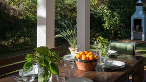 Un área de comedor al aire libre con una mesa de madera dispuesta para una comida, rodeada de columnas blancas y con vistas a un jardín con árboles y una pequeña construcción.