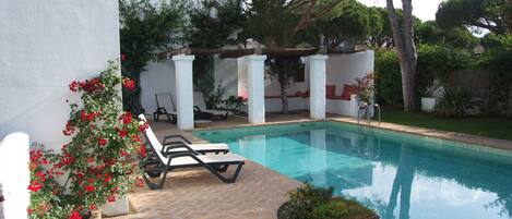 A swimming pool with a tiled deck, lounge chairs, and a pergola with red cushions.