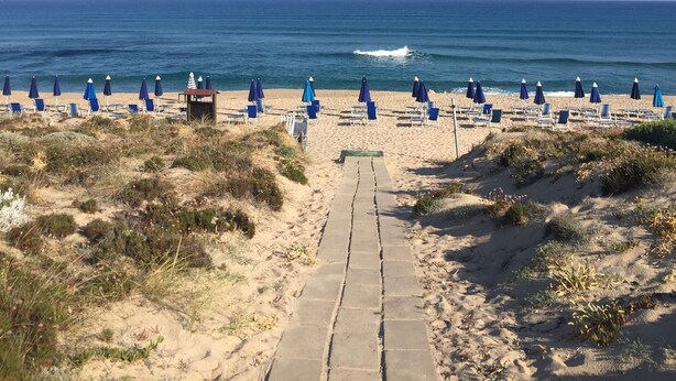 A beach with a concrete pathway leading to the sea, lined with blue beach chairs and umbrellas. - directly on the sea new apartment with garden and swimming pool (badesi)