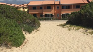 A sandy pathway leading to a row of orange buildings with a tiled roof, surrounded by greenery. - directly on the sea new apartment with garden and swimming pool (badesi)