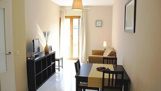 A compact living space with a dining table, chairs, a sofa, a TV on a black stand, and a lamp.