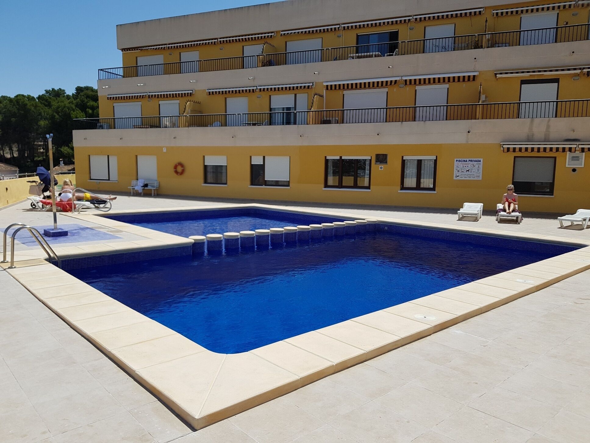 A swimming pool with a tiled deck, surrounded by lounge chairs and a building with balconies.