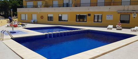 A swimming pool with a tiled deck, surrounded by lounge chairs and a building with balconies.