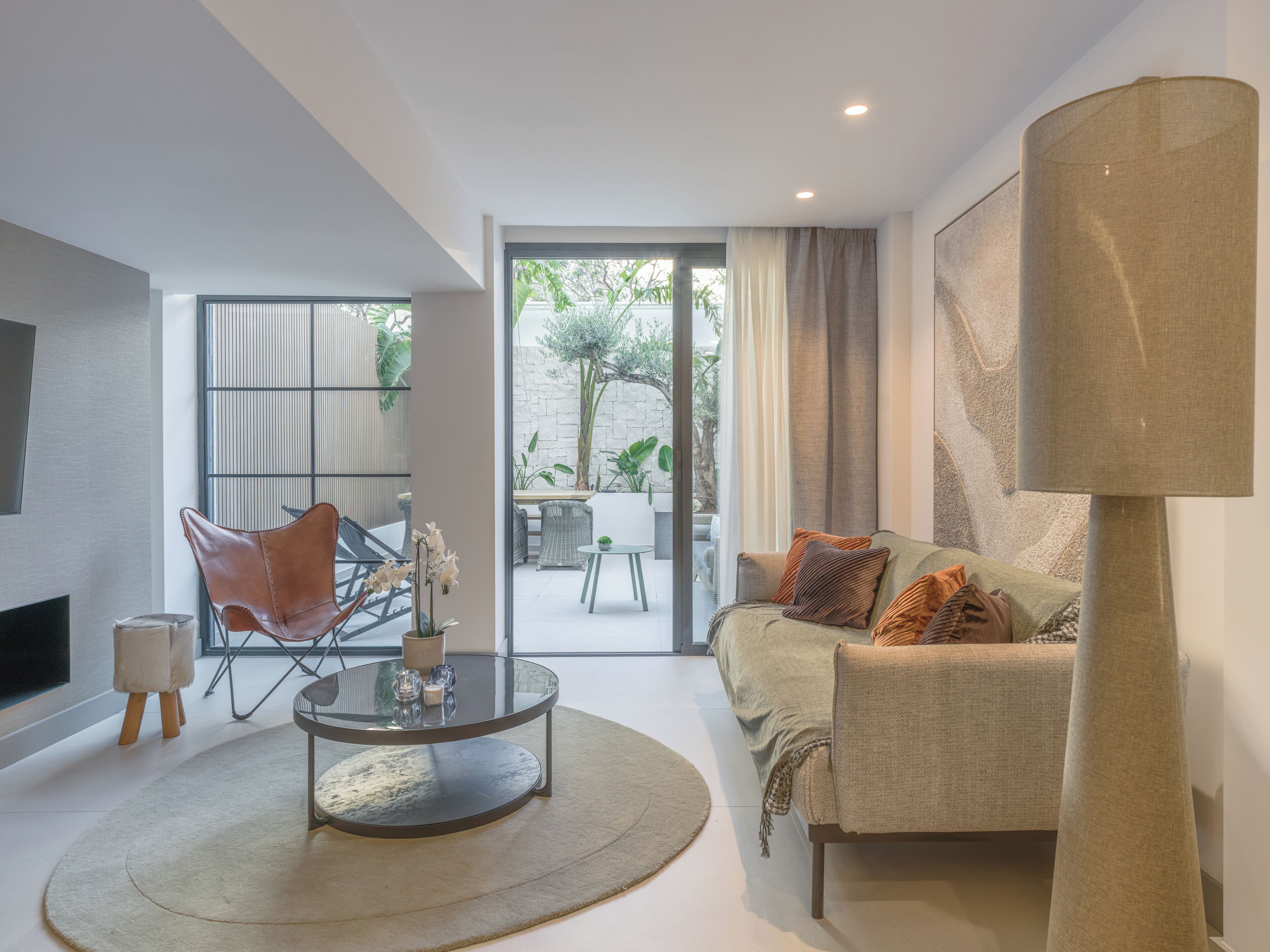 Een moderne woonkamer met een glazen salontafel, een bank met kussens, een grote lamp en uitzicht op een terras.