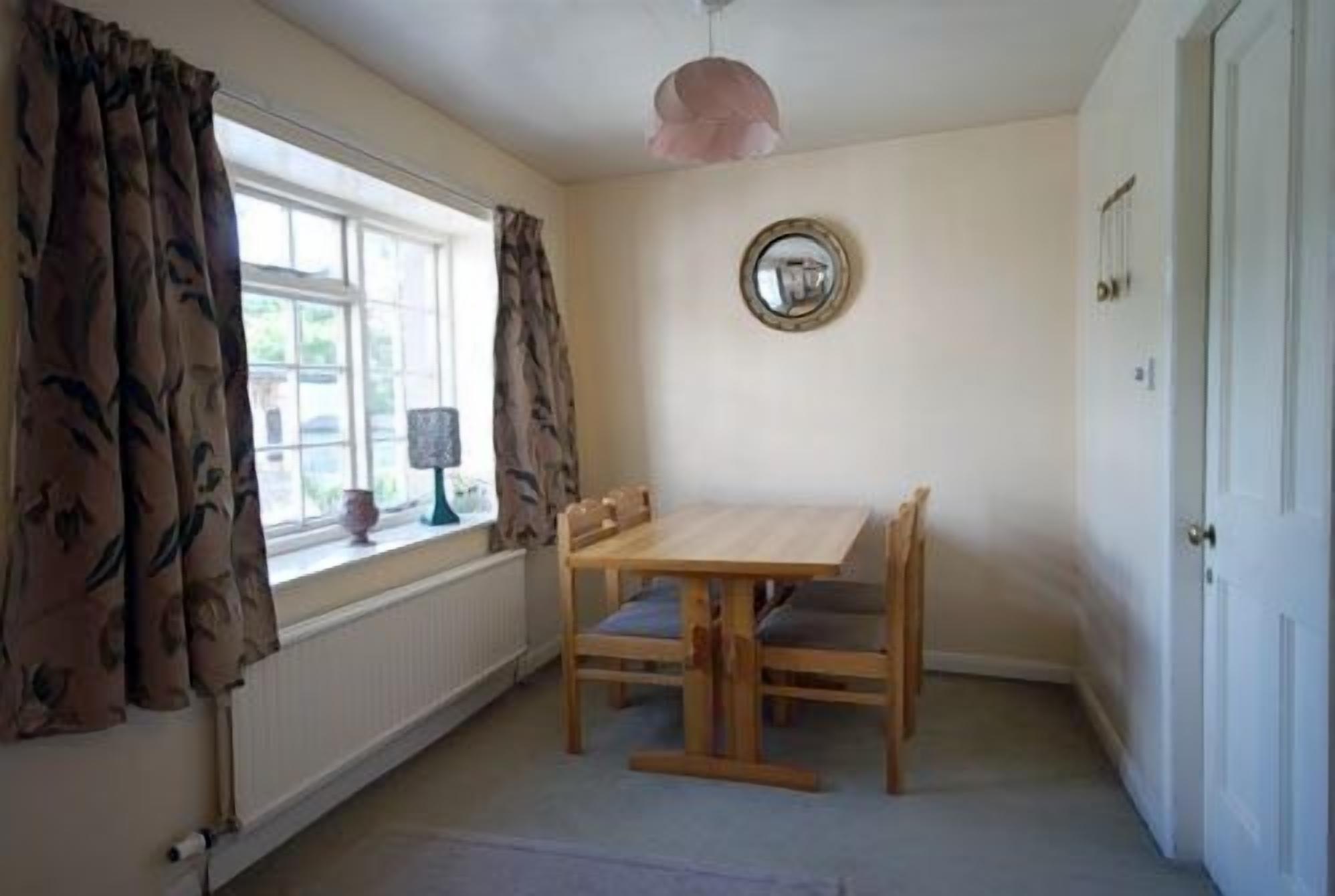 A room with a wooden table and chairs, a window with curtains, a wall-mounted mirror, and a radiator.