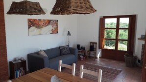 A living room with a sofa, a dining table, a painting on the wall, a lamp, and a window with a view of greenery.