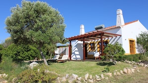 A white building with a red-tiled roof, a pergola with chairs, and a large tree in the foreground.