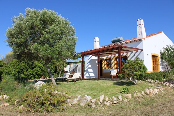A white building with a red-tiled roof, a pergola with chairs, and a large tree in the foreground.