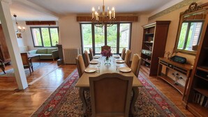 A dining room with a large wooden table set for six, surrounded by chairs. There is a chandelier above the table, and a large window with a view of greenery outside. - Sarlat La Caneda: SPACIOUS APARTMENT WITH GARDEN AND PRIVATE SWIMMING POOL (Sarlat-la-Canéda)