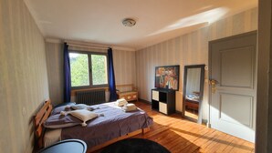 A bedroom with a bed, a dresser, a chair, a radiator, a window with curtains, a door, and a painting on the wall. - Sarlat La Caneda: SPACIOUS APARTMENT WITH GARDEN AND PRIVATE SWIMMING POOL (Sarlat-la-Canéda)