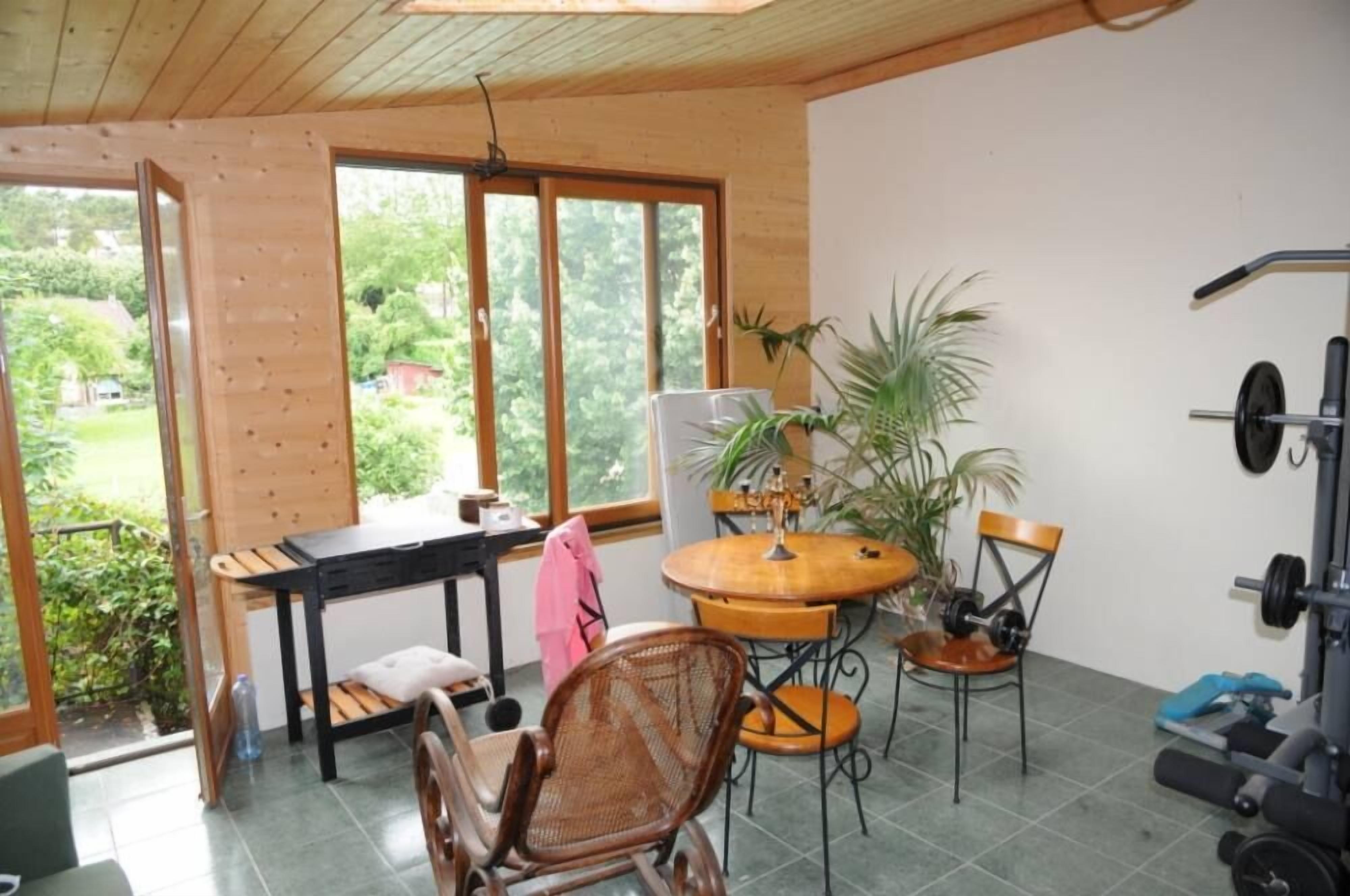 Une pièce avec un plafond en bois, de grandes fenêtres, une table avec des chaises, une plante et du matériel de musculation.