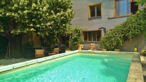 A swimming pool with a wooden deck and chairs, surrounded by a house and greenery. - Sarlat La Caneda: SPACIOUS APARTMENT WITH GARDEN AND PRIVATE SWIMMING POOL (Sarlat-la-Canéda)