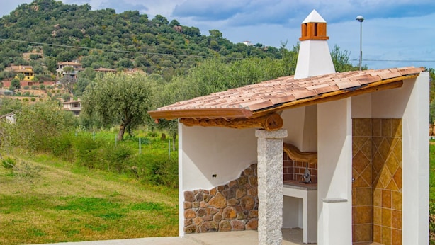 Uma pequena cozinha ao ar livre com parede de pedra e telhado ladrilhado, situada em uma encosta com casas ao fundo.